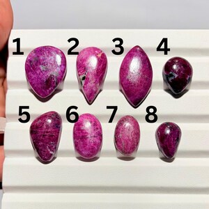 May include: An assortment of eight polished, teardrop and oval-shaped gemstones in varying sizes. The stones are a deep magenta color with lighter pink and white veining. The gemstones are arranged on a white surface.