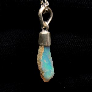 Puede incluir: Un collar de plata con un colgante de ópalo en bruto. El ópalo tiene una textura natural y rugosa con destellos iridiscentes azules y verdes. El colgante está unido a una cadena de plata, creando una joya sencilla pero elegante.