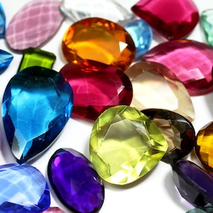 Op de afbeelding: Een verzameling gefacetteerde edelstenen in verschillende vormen en kleuren, waaronder blauw, roze, geel, oranje en paars. De edelstenen zijn verspreid op een witte ondergrond, wat hun helderheid en glans benadrukt.