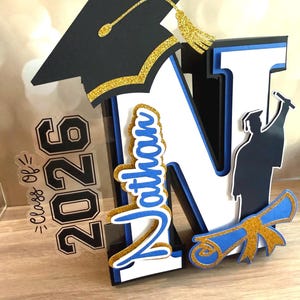 May include: A graduation cake topper with a large letter "N" in white, black, and blue, with the name "Nathan" in blue script. Features a graduation cap, diploma, and the year "2026". Decorated with gold glitter accents.