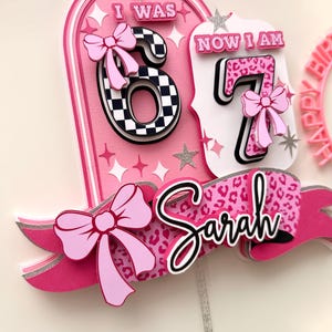 Puede incluir: Adorno para tarta de cumpleaños rosa y blanco con el texto "I WAS 6 NOW I AM 7" y el nombre "Sarah". El número 6 tiene un patrón de cuadros y el número 7 un estampado de leopardo. El adorno tiene lazos rosas y estrellas.