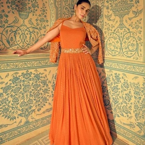 May include: An orange formal gown with a matching sequined jacket. The dress has a fitted bodice, a pleated skirt, and a jeweled belt. The jacket is draped over the shoulders. The background has a floral pattern.
