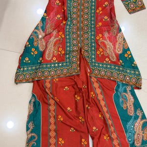 May include: A two-piece outfit featuring a long-sleeved tunic and wide-leg pants. The ensemble is primarily red with teal and gold accents. The tunic has a collared neckline and is adorned with floral and paisley patterns. The pants mirror the design, creating a cohesive look.