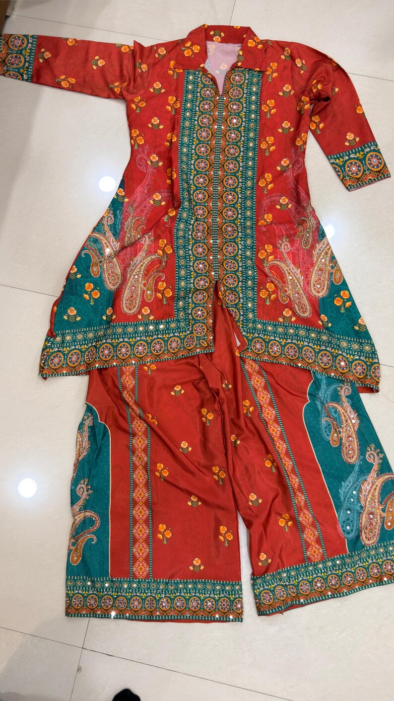 May include: A two-piece outfit featuring a long-sleeved tunic and wide-leg pants. The ensemble is primarily red with teal and gold accents. The tunic has a collared neckline and is adorned with floral and paisley patterns. The pants mirror the design, creating a cohesive look.