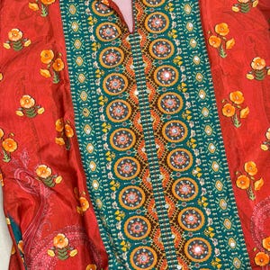May include: A red collared shirt with a floral and geometric pattern. The shirt features a central panel of teal and gold geometric designs, bordered by red fabric with yellow floral accents. The collar and sleeves also have floral patterns.