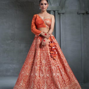 May include: A vibrant orange and gold lehenga, featuring a one-shoulder top and a detailed skirt. The skirt showcases a floral and peacock pattern. The top has one long sleeve and a decorative floral design. A cluster of orange fabric adorns the waist.