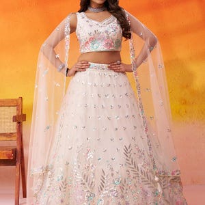 May include: A cream-colored, sleeveless lehenga with floral embroidery and sequin accents. The ensemble includes a matching cropped top and a sheer, flowing dupatta. The skirt features detailed floral patterns in pink, blue, and silver.