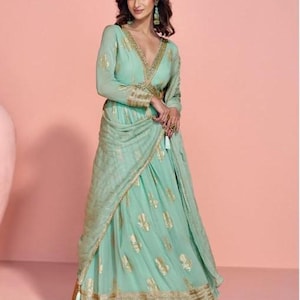 Puede incluir: Un vestido formal verde menta con detalles dorados. El vestido presenta un corpiño cruzado con escote pronunciado y mangas largas. La falda es de corte A con estampados de hojas doradas. El vestido está adornado con detalles dorados.