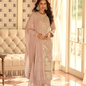 以下が含まれることがあります： 薄いピンク色のサルワールカミーズのアンサンブル。チュニックとパンツには金色の刺繍が施され、透明なドゥパッタが付いています。金色のイヤリングとサンダルを合わせています。特別な機会に適した衣装です。