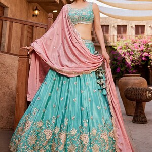May include: A turquoise and gold embroidered lehenga choli set. The set includes a sleeveless top with intricate embroidery, a flowing skirt, and a pink dupatta. The outfit is embellished with sequins and gold thread.