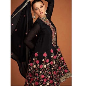 May include: A black embroidered dress with a matching sheer scarf. The dress features a floral pattern in pink and white, with gold accents on the sleeves and hem. The model is wearing matching earrings and a necklace.