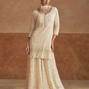 Op de afbeelding: Crèmegekleurde geborduurde salwar kameez set. De set bestaat uit een lange tuniek met een geometrisch patroon, driekwartmouwen en een bijpassende broek met wijde pijpen. De outfit wordt gecompleteerd met een sjaal en is versierd met ingewikkelde borduursels.