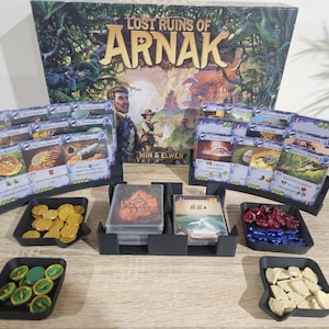May include: The board game "Lost Ruins of Arnak" is displayed with game cards, tokens, and a box. The game components are organised in black trays. The box art features a jungle scene with the game title and the names "Min & Elwen".