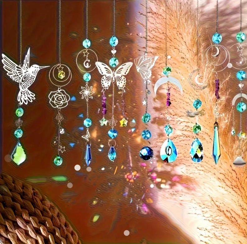 May include: A collection of hanging suncatchers featuring metal silhouettes of hummingbirds, butterflies, moons, and stars. Each piece is accented with colorful faceted glass beads and crystals in shades of blue, green, and purple, suspended on thin metal chains.