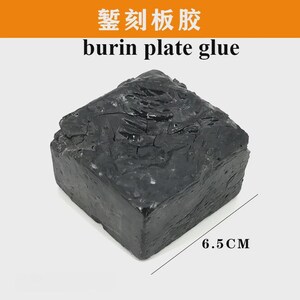 May include: A dark gray, rectangular block of burin plate glue. The block has a rough, textured surface and measures 6.5 cm in length. The text "burin plate glue" is printed above the block.
