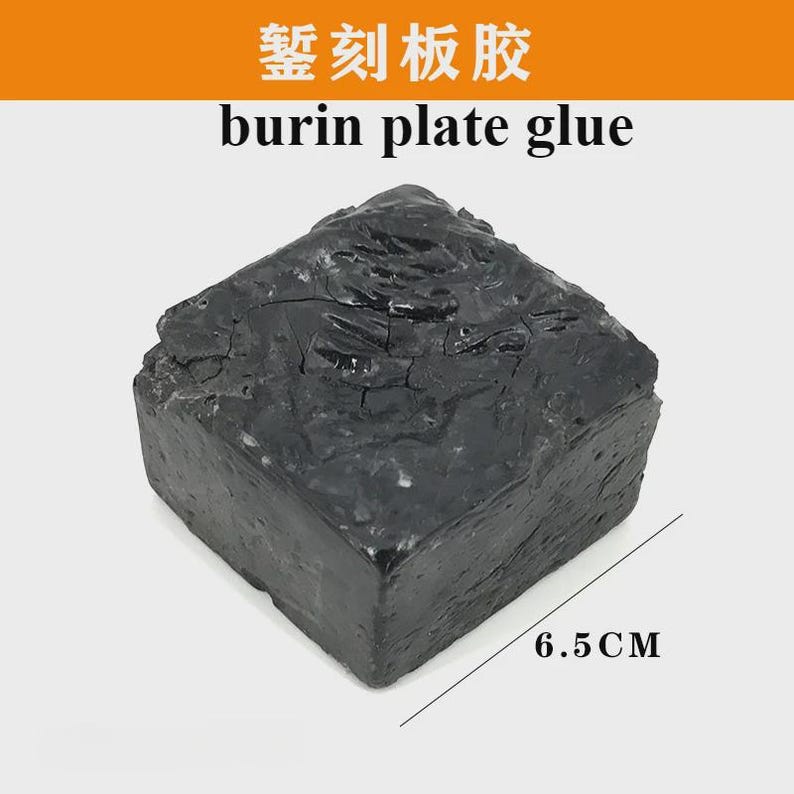 May include: A dark gray, rectangular block of burin plate glue. The block has a rough, textured surface and measures 6.5 cm in length. The text "burin plate glue" is printed above the block.