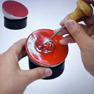 May include: A red wax seal with a silver ring being set into it. A wooden handled tool is used to set the ring. A second, similar seal is in the background. The seals are on a black base.