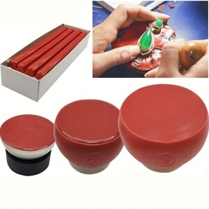 May include: A collection of red sealing wax sticks in a white box, alongside three red wax seal stamps with white and black bases. The image also shows a person working on a piece of jewelry.