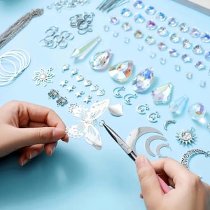 May include: A jewelry making kit featuring silver-toned metal charms in butterfly, sun, and moon shapes, alongside iridescent crystal beads and jump rings. A pair of pliers is used to attach a jump ring to a silver butterfly charm on a light blue surface.