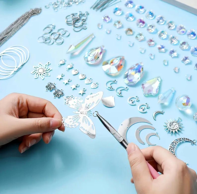 May include: A jewelry making kit featuring silver-toned metal charms in butterfly, sun, and moon shapes, alongside iridescent crystal beads and jump rings. A pair of pliers is used to attach a jump ring to a silver butterfly charm on a light blue surface.