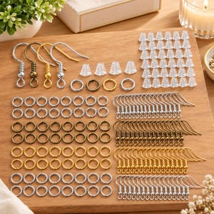 May include: A jewelry making kit containing silver, gold, and bronze tone earring hooks, numerous circular jump rings in matching metallic finishes, and a large collection of clear silicone earring backs arranged on a wooden surface.
