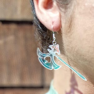 May include: A silver earring shaped like a battle axe with a heart-shaped cutout. The axe head is teal and the handle is clear. The earring is hanging from a silver hook.