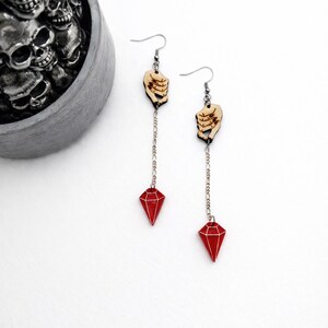 May include: A pair of dangle earrings featuring a wooden hand design holding a silver chain that leads to a red diamond-shaped charm. The earrings are set against a white background, with a decorative skull container in the background.