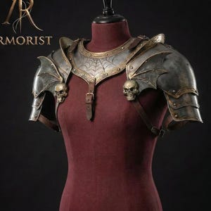 Puede incluir: Una detallada pieza de armadura artesanal exhibida en un maniquí. La armadura del hombro es metálica con diseños de alas de murciélago, detalles de calaveras y un patrón de telaraña. La armadura se asegura con correas y hebillas de cuero. La palabra "ARMORIST" es visible.