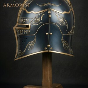 K&ouml;nnte beinhalten: Dunkelblauer und goldener mittelalterlicher Helm mit aufwendigen goldenen Details und dem Wort "ARMORIST" in der oberen linken Ecke. Der Helm wird auf einem Holzst&auml;nder vor dunklem Hintergrund ausgestellt.