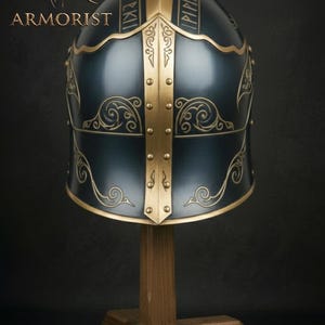 K&ouml;nnte beinhalten: Dunkelblauer Helm mit goldenen Akzenten und dekorativen Schn&ouml;rkeln, pr&auml;sentiert auf einem Holzst&auml;nder. Der Helm hat goldene Verzierungen und Nieten, mit Text oben. Das Wort "ARMORIST" ist in goldenen Buchstaben zu sehen.
