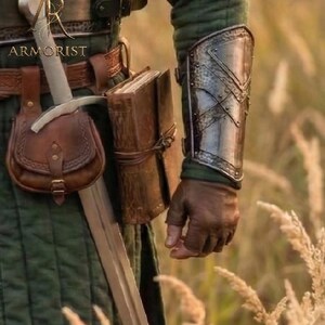 K&ouml;nnte beinhalten: Ein mittelalterliches Ensemble mit einer Ledertasche, einem Schwert und einem Metallarmschutz. Das Bild zeigt auch ein ledergebundenes Buch und das Wort "ARMORIST". Das Outfit ist vor dem Hintergrund von hohem Gras platziert.