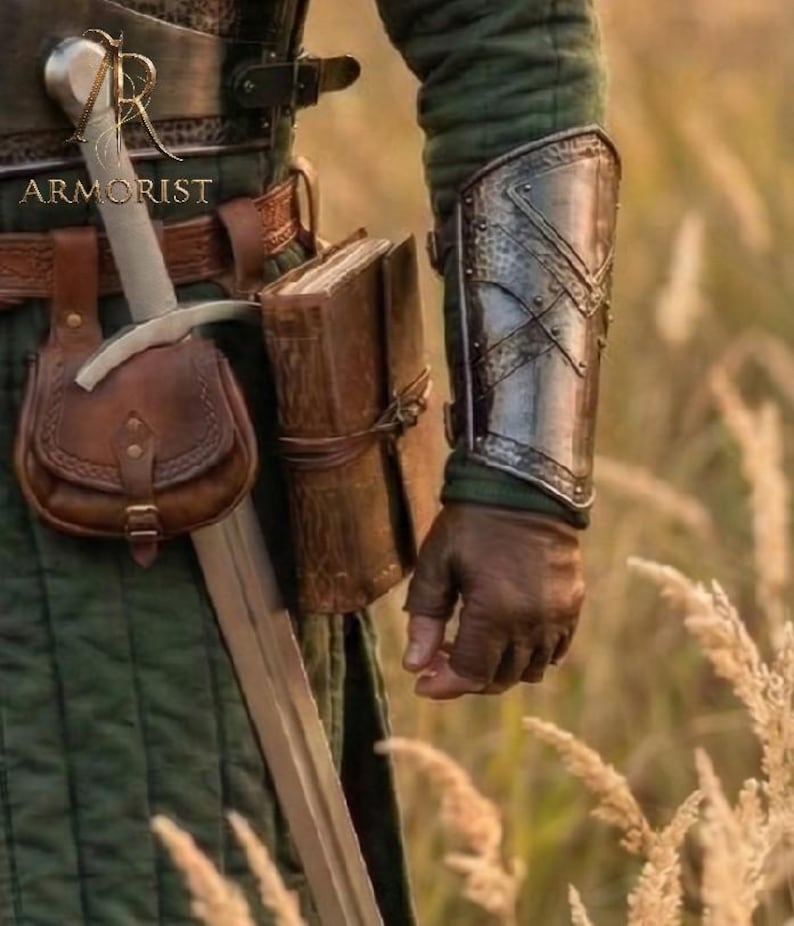 K&ouml;nnte beinhalten: Ein mittelalterliches Ensemble mit einer Ledertasche, einem Schwert und einem Metallarmschutz. Das Bild zeigt auch ein ledergebundenes Buch und das Wort "ARMORIST". Das Outfit ist vor dem Hintergrund von hohem Gras platziert.
