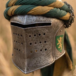K&ouml;nnte beinhalten: Ein mittelalterlicher Stahlhelm mit genieteter Konstruktion und zahlreichen Bel&uuml;ftungsl&ouml;chern. Der Helm hat eine gr&uuml;n-goldene Stoffwicklung und ein Schildemblem. Das Wort "ARMORIST" ist im Bild sichtbar.