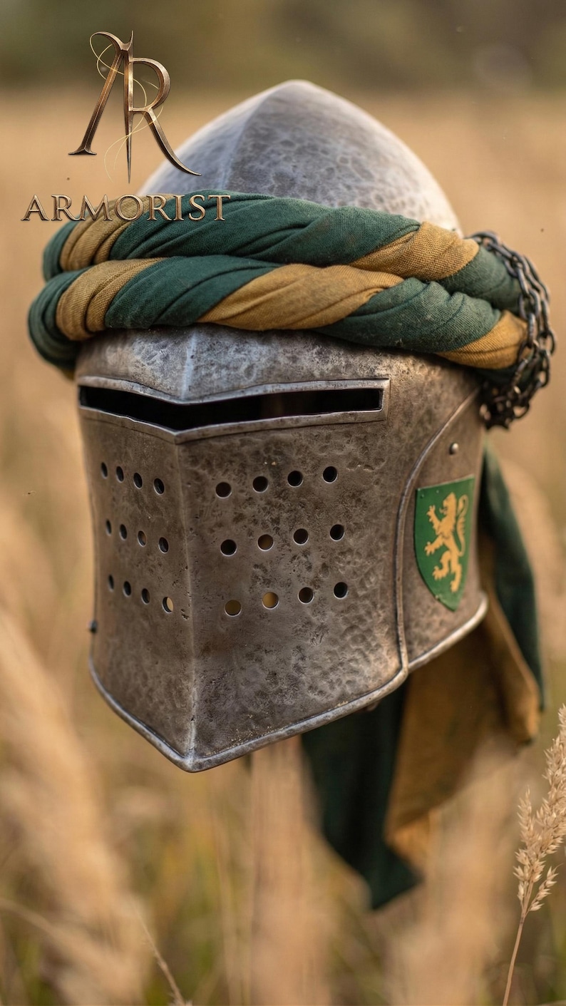 K&ouml;nnte beinhalten: Ein mittelalterlicher Stahlhelm mit genieteter Konstruktion und zahlreichen Bel&uuml;ftungsl&ouml;chern. Der Helm hat eine gr&uuml;n-goldene Stoffwicklung und ein Schildemblem. Das Wort "ARMORIST" ist im Bild sichtbar.