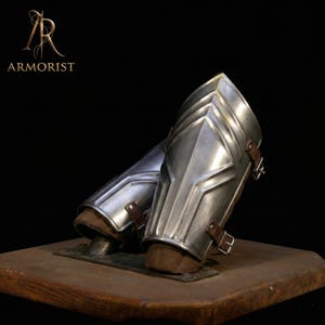 Pode incluir: Par de protetores de braço de metal prateado com correias e fivelas de couro castanho. Os protetores de braço são exibidos em uma base de madeira. A palavra "ARMORIST" está impressa em texto preto.