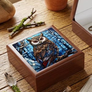 Peut inclure: Une boîte à bijoux en bois avec une illustration détaillée d'un hibou sur le couvercle. Le hibou est représenté avec des tons bruns, bleus et rouges, perché sur une branche sur fond bleu. La boîte est rectangulaire avec une finition en bois foncé.