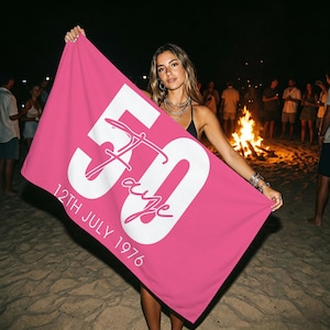 May include: A bright pink rectangular towel featuring a large white number 50 with a stylized script name overlaying the center. Below the number, the text 12TH JULY 1976 is printed in white capital letters. The towel has a smooth, flat fabric texture.