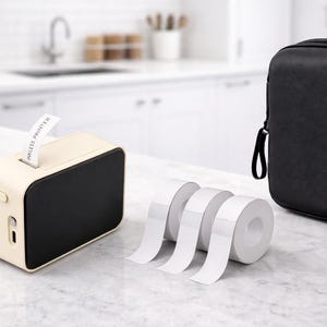 Puede incluir: Una impresora de etiquetas inalámbrica crema y negra con una etiqueta que se extiende desde la parte superior. También hay rollos de cinta de etiquetas blanca y un estuche de transporte negro. La etiqueta dice "WIRELESS PRINTER".