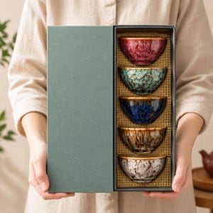 Pode incluir: Um conjunto de cinco tigelas de chá de cerâmica numa caixa de presente. As tigelas são em tons de vermelho, verde-azulado, azul, castanho e branco. Cada tigela tem um esmalte salpicado único e uma borda dourada. A caixa de presente é verde suave.