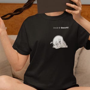 May include: Black t-shirt with a white anime-style graphic and the text "I'm B-B-Bocchi!". The shirt is short-sleeved and appears to be made of a soft material. The shirt is being worn by a person.