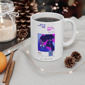 Puede incluir: Taza de cerámica blanca con una bebida negra en su interior. La taza presenta un gráfico colorido con el texto "BABY, I'M dancin' in the DARK" y "WITH YOU between my ARMS". El gráfico incluye notas musicales y la palabra "Perfect".