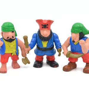 May include: Three colorful pirate figurines. The figures are wearing red, blue, and yellow clothing, with green and red hats. One figure holds an axe, another a sword, and the third a club. The figures have black beards and mustaches.