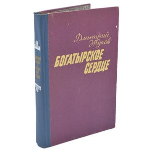 May include: A hardcover book with a blue spine and a maroon cover. The cover features gold lettering that reads "Дмитрий Жуков БОГАТЫРСКОЕ СЕРДЦЕ". The book is slightly angled, showcasing its title and author.