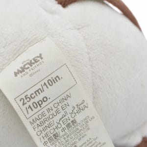May include: A white plush toy with a tag that reads "MICKEY AND FRIENDS" and "25cm/10in." The tag also indicates the toy was made in China. The toy has a soft, textured appearance.