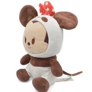 May include: A plush toy of a mouse character wearing a white hooded outfit with brown spots and a red bow with white polka dots. The toy has large brown ears and a brown tail. The mouse is sitting down.