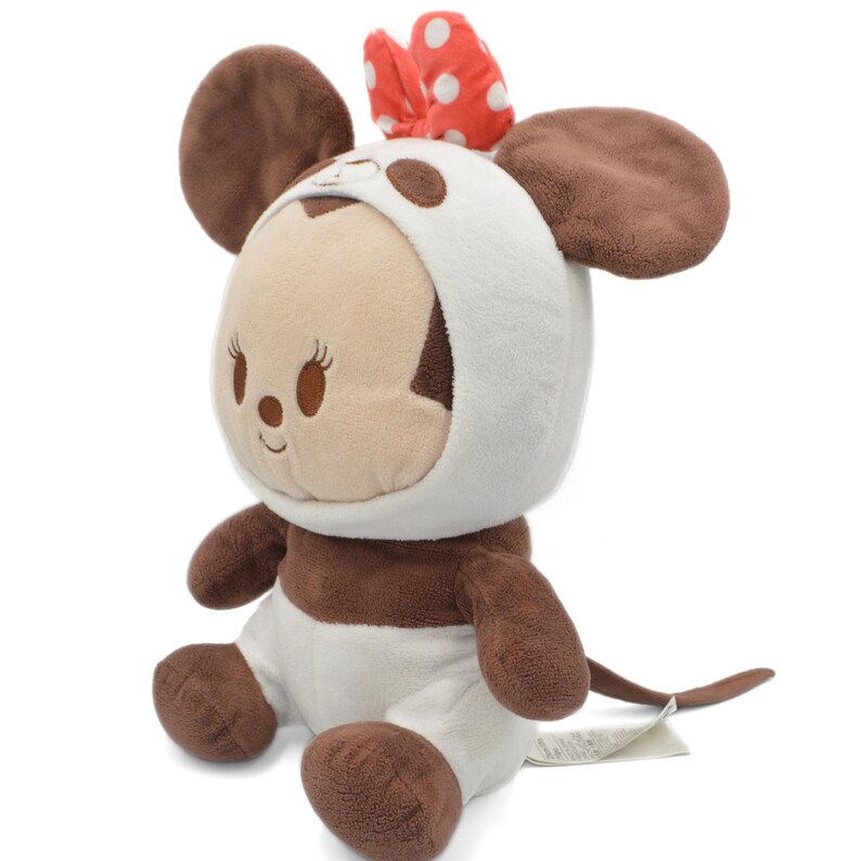 May include: A plush toy of a mouse character wearing a white hooded outfit with brown spots and a red bow with white polka dots. The toy has large brown ears and a brown tail. The mouse is sitting down.