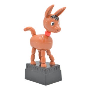 May include: A brown plastic donkey figurine with a red collar, black hair detail, and white eyes. The donkey stands on a rectangular black base with a textured grass-like pattern along the top edge, featuring articulated legs and a small tail.