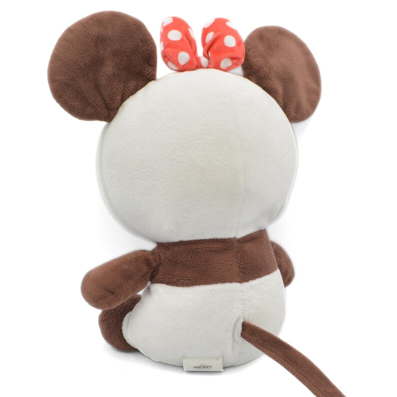 May include: A plush toy mouse with brown ears, arms, and tail, and a white body. The mouse has a red bow with white polka dots on its head. The toy is sitting down, with its back facing the viewer.