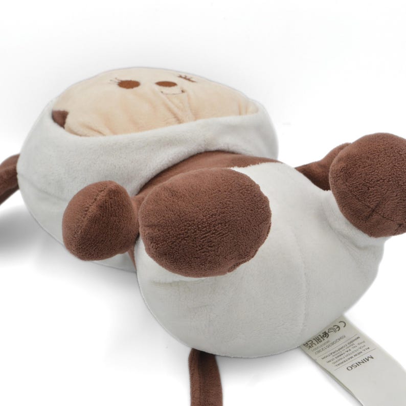 May include: A plush toy cow in a lying position, featuring a white body with brown spots and limbs. The cow has a beige head with embroidered eyes and a smiling mouth. The toy has a soft, fuzzy texture and a tag that reads "MINISO".