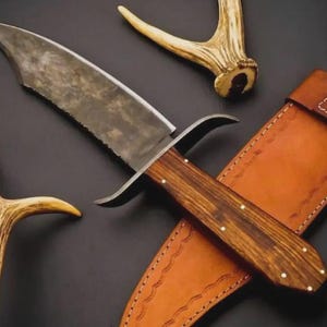 May include: A fixed-blade knife with a dark, textured steel blade, a wooden handle secured with small brass pins, and a curved metal guard. The knife is accompanied by a tan leather sheath featuring decorative stitching and a brass snap closure.